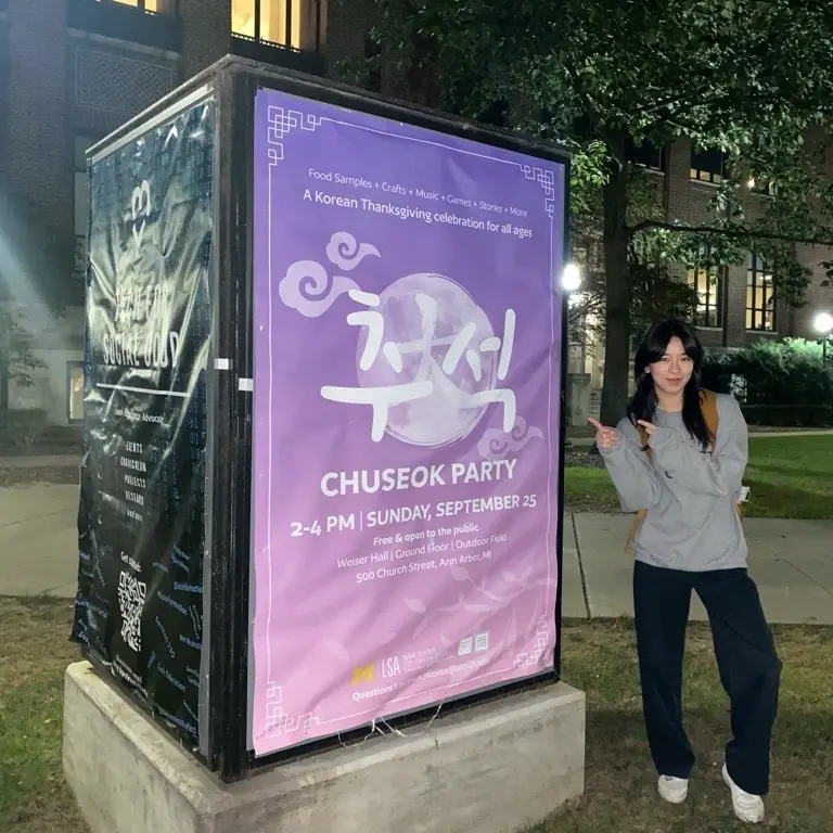 Purple Chuseok Party poster for a Korean Thanksgiving celebration, displayed on a campus kiosk at night, with a student pointing to it